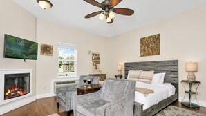 A bedroom with a fireplace, a bed with a wooden headboard, two armchairs, a ceiling fan, and artwork on the walls.