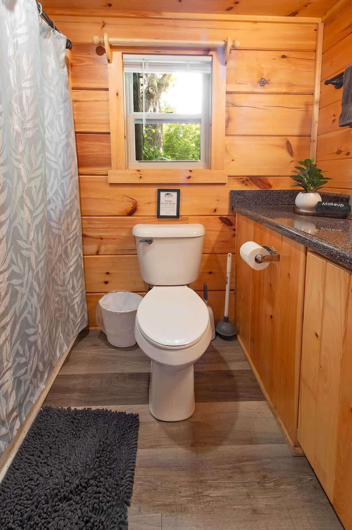 Baño de una cabaña de madera pequeña con inodoro, ventana, lavamanos y cortina de ducha.