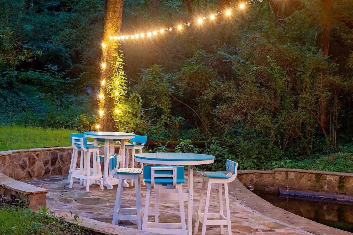 Une terrasse extérieure avec des chaises blanches et bleues, une table en pierre et des guirlandes lumineuses.