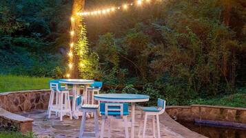 Une terrasse extérieure avec des chaises blanches et bleues, une table en pierre et des guirlandes lumineuses.