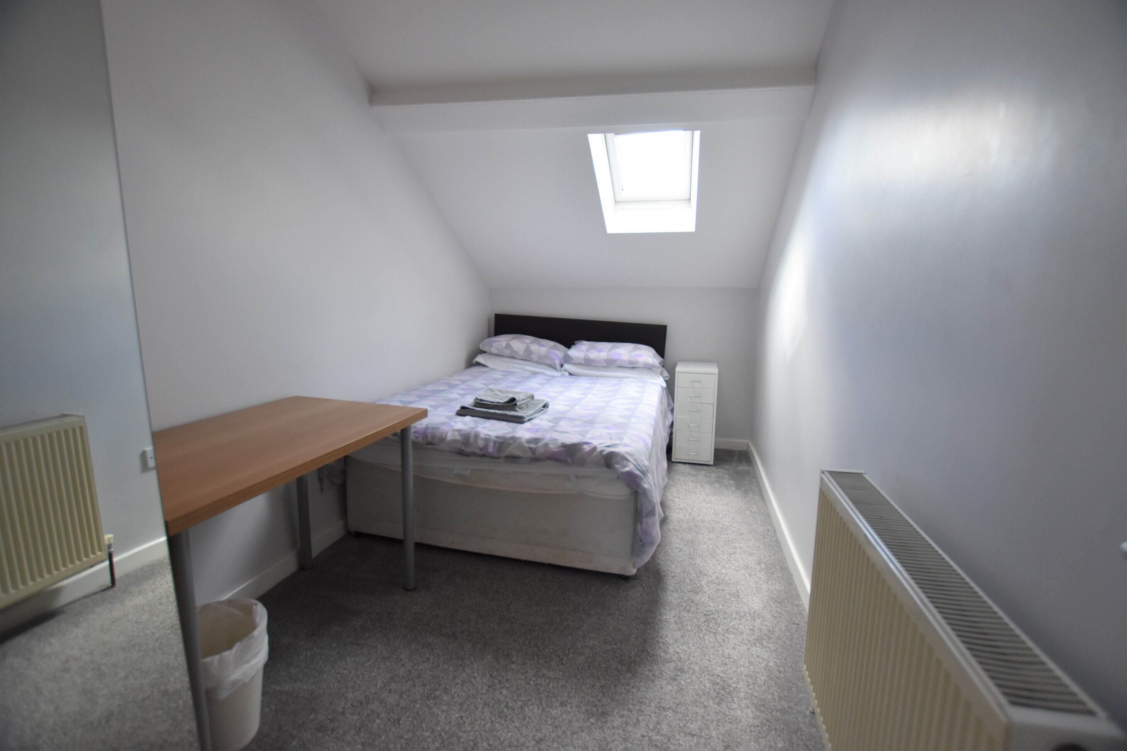 A small, neatly arranged bedroom with a single bed, a small table, and a window providing natural light.
