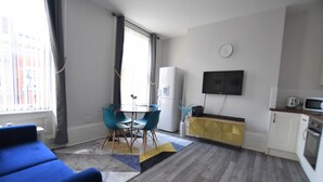 Ein modernes Wohnzimmer mit einem blauen Sofa, einem Esstisch mit Stühlen, einer Küchenzeile mit Fernseher und Kühlschrank.