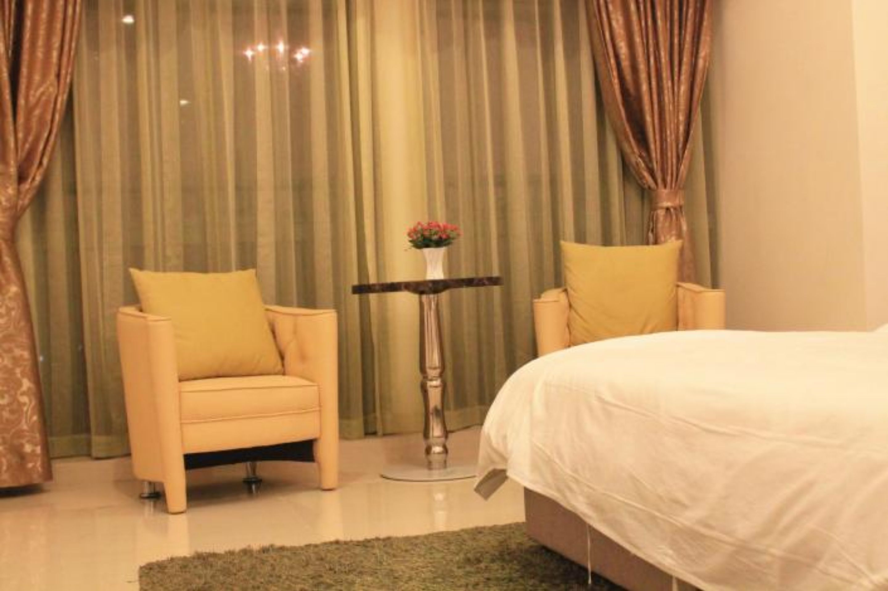 A bedroom with a bed, two chairs, a small table with a vase, and curtains.