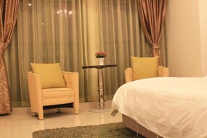 Un dormitorio con una cama, dos sillas, una mesita de noche con un florero y cortinas.