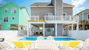 A pool area with white and yellow lounge chairs, a covered deck, and a two-story building with a balcony. - Tar Healin': Private Saltwater Pool and Ocean Views on Carolina Coast (Oak Island)