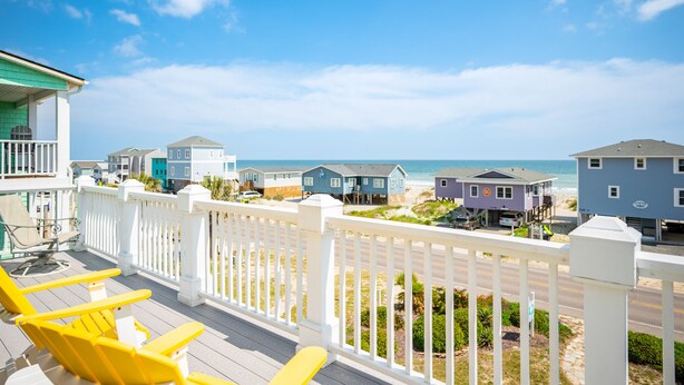 A coastal view with a wooden deck, yellow chairs, and a white fence overlooking beach houses and the ocean. - Tar Healin': Private Saltwater Pool and Ocean Views on Carolina Coast (Oak Island)