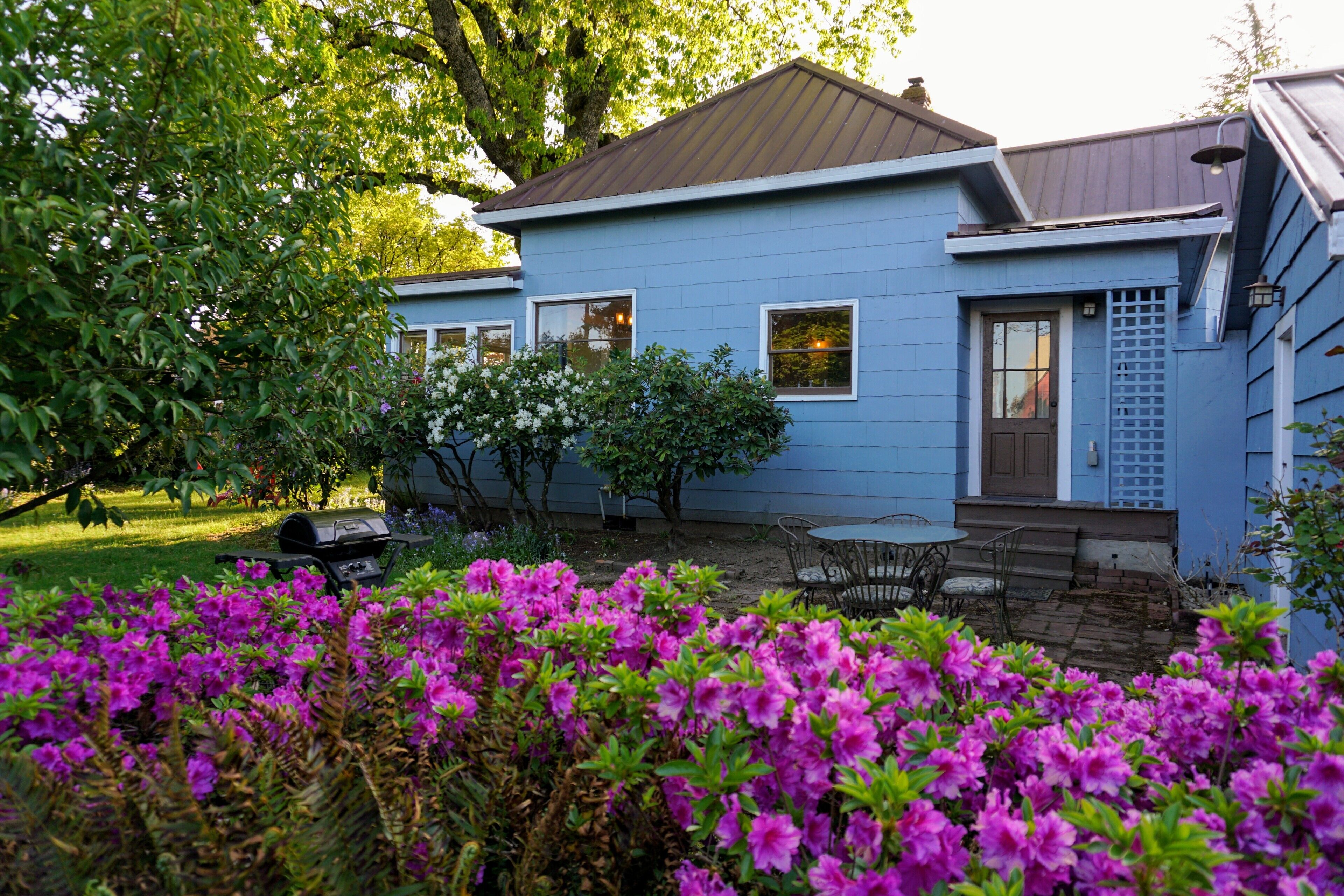 A blue house with a front porch, a grill, and a garden with purple flowers.