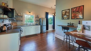 A modern kitchen with white cabinets, stainless steel appliances, and a wooden countertop. There is a dining area with a table and chairs, and a window with curtains.