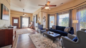 A spacious living room with a dark blue sofa, a grey armchair, and a wooden coffee table. A ceiling fan and a chandelier are visible.