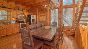 A wooden cabin interior with a dining table, chairs, a chandelier, and a kitchen area with appliances. - MAJESTIC MEMORIES - 3 LEVEL CUSTOM CABIN! (Sevierville)