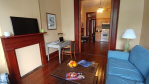 A living room with a blue sofa, a coffee table with magazines and fruit, a TV on a stand, and a kitchen in the background.