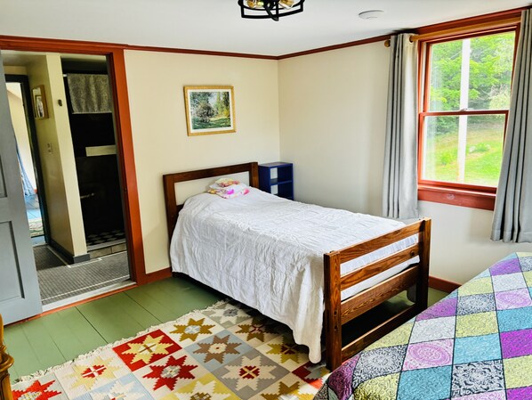 A bedroom with a single bed, a window with curtains, a door leading to a hallway, and a small blue cabinet.