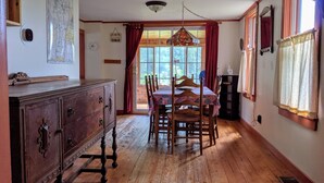 A dining area with a wooden table and chairs, a vintage dresser, and a large window with curtains.