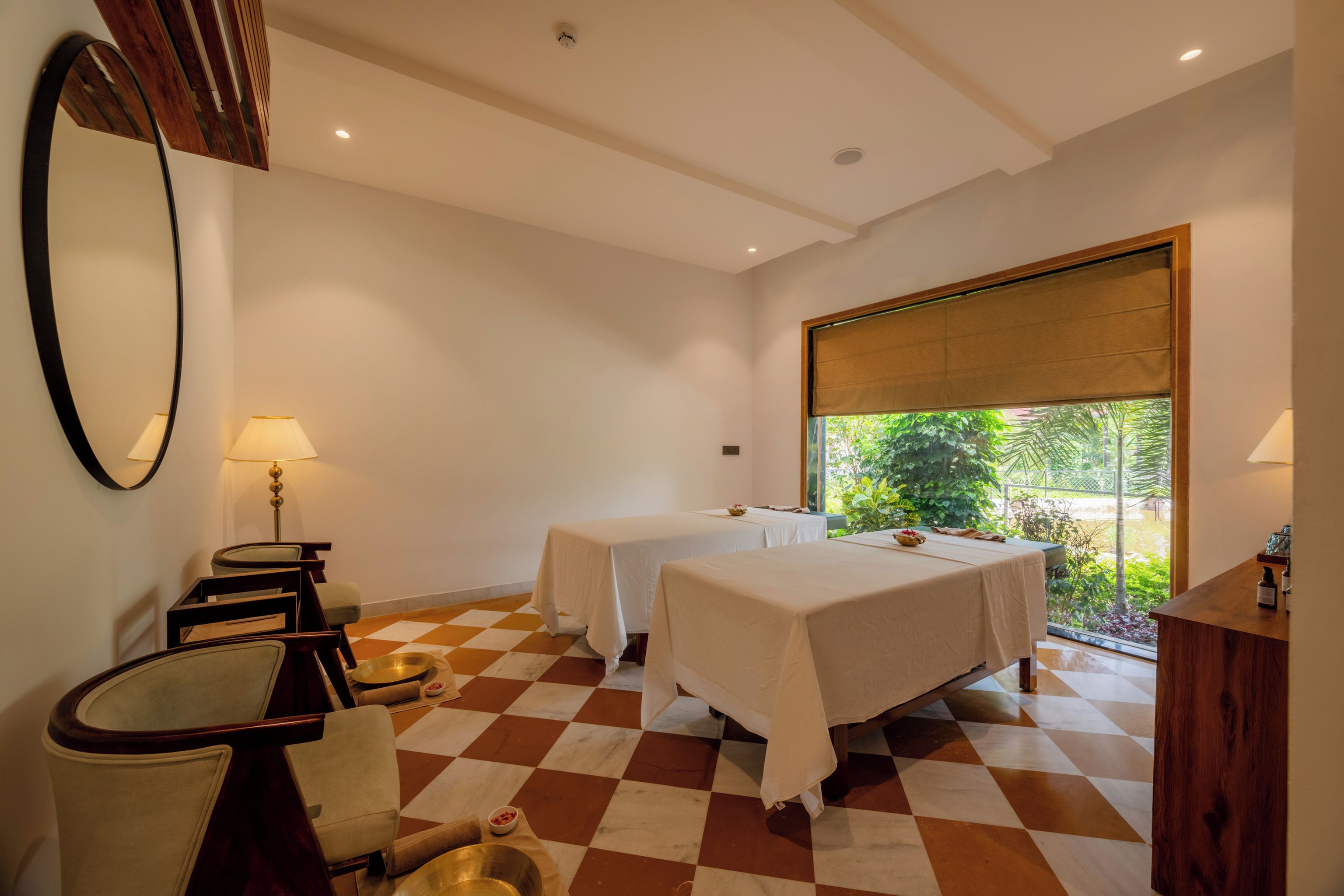 Une chambre d’hôtel avec deux lits, un sol à carreaux, une grande fenêtre donnant sur un paysage verdoyant, un bureau avec une lampe et un miroir au mur.