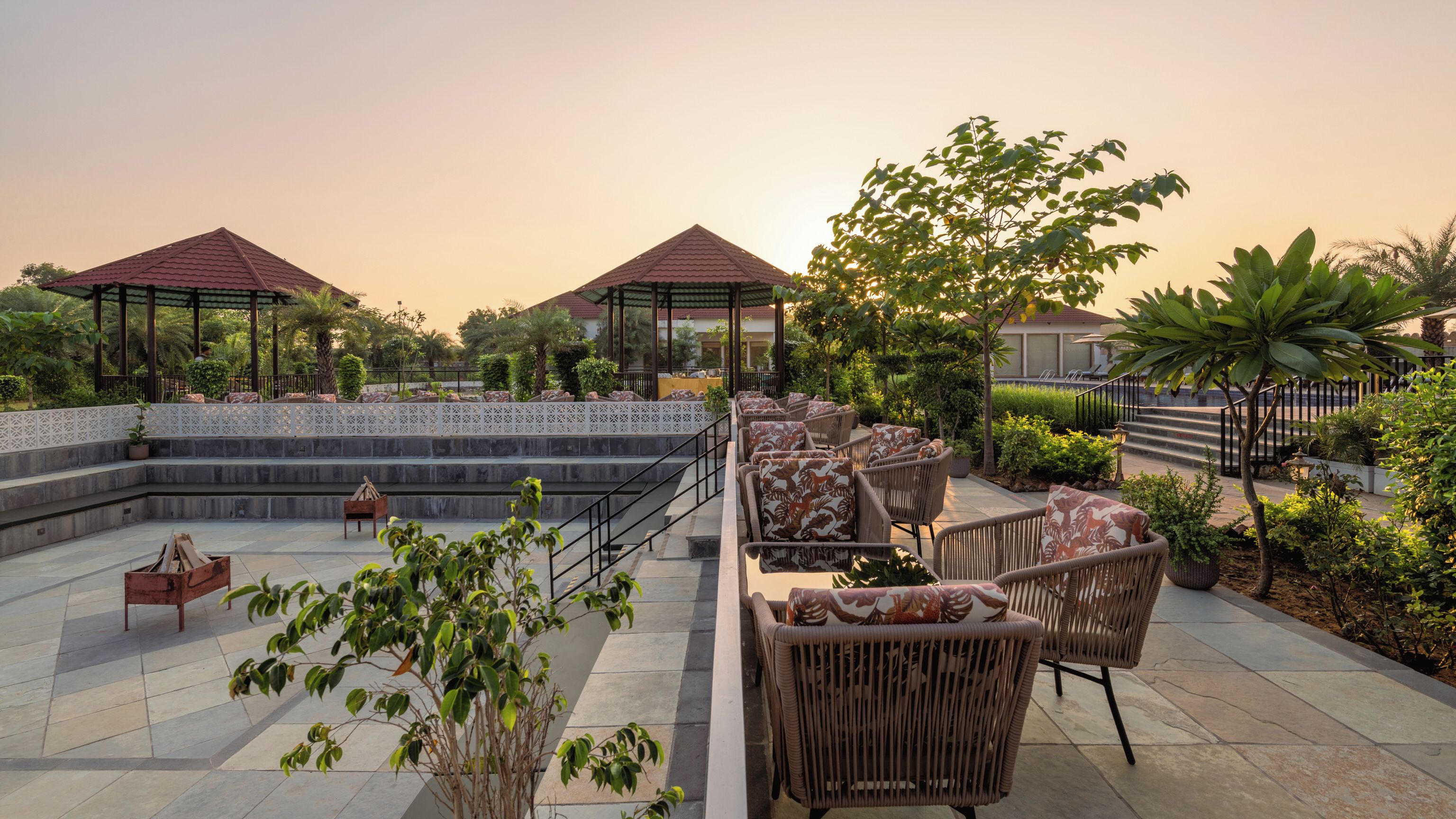 An outdoor seating area with wicker furniture and a stone floor, surrounded by greenery and a gazebo structure.