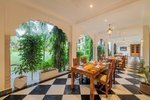 Un comedor de hotel con piso a cuadros, mesas y sillas de madera, ventanas amplias con follaje afuera y vista de un edificio al fondo.