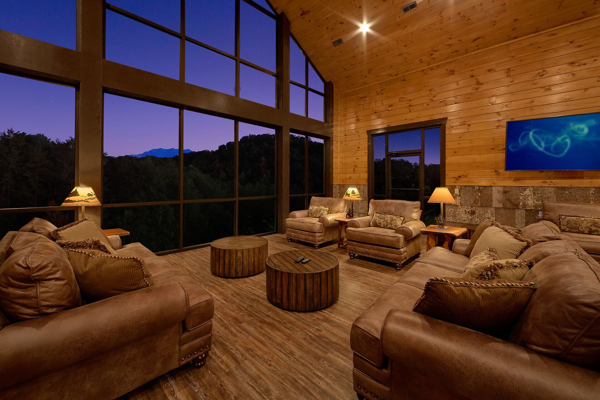 A spacious living room with leather sofas and armchairs, large windows offering a view of the outdoors, and a wooden ceiling.