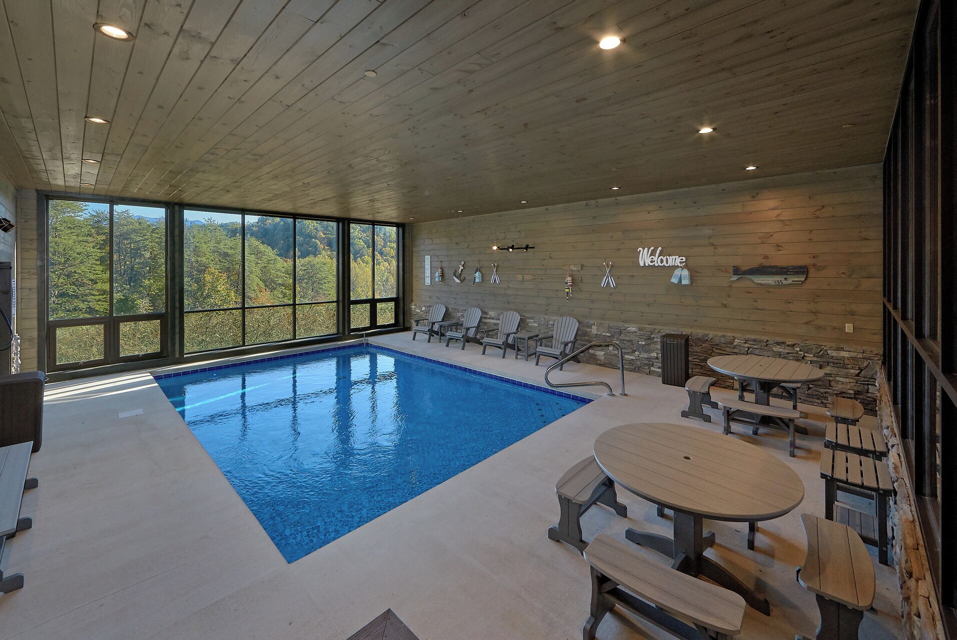 An indoor swimming pool with a view of trees, wooden deck chairs, and a stone wall with a 'Welcome' sign.