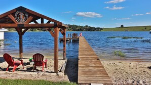 A wooden pier leading to a gazebo with a Corona Corona logo, red chairs, and a view of a lake with boats and a distant hill.