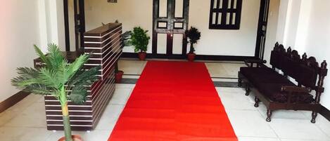A hotel lobby with a red carpet, a reception desk, a bench, and potted plants.