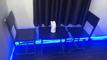 A room with a dark wooden floor, a black table with a white object on it, and two black chairs. Blue LED lighting is along the baseboard.