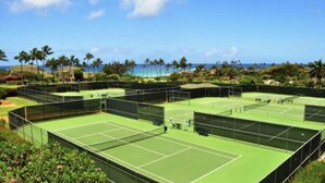 Tennisanlæg med flere baner, omgivet af grønt og palmer, med udsigt til havet.