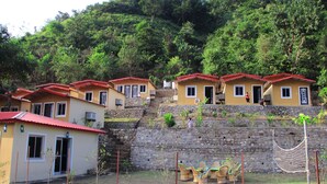 A row of yellow buildings with red roofs, each with a door and windows, situated on a hillside with a stone wall in the background. - Panchvati cottage (Narendranagar)