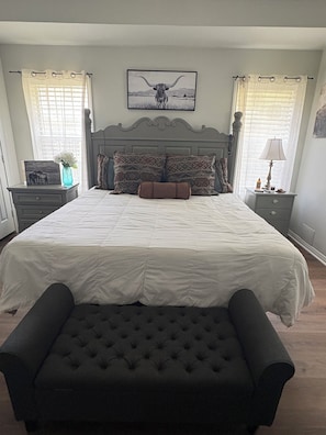A bedroom with a large bed, a headboard, a bedside table, a lamp, a nightstand, and a window with curtains.