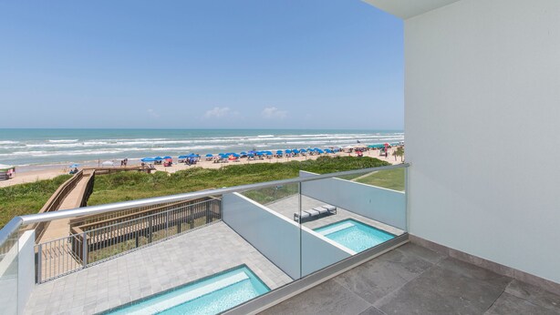 Un balcon avec une piscine donnant sur une plage parsemée de parasols et où se trouvent des gens.