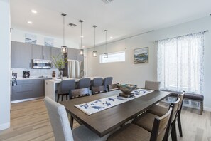 Ett modernt kök med en central köksö, en matplats med ett långt bord och ett stort fönster med gardiner.