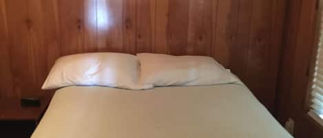 A room with wooden panel walls, a bed with white bedding, and a window with blinds.