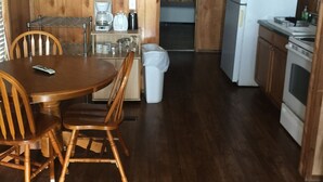 A wooden interior with a dining table and chairs, a kitchen area with appliances, and a trash bin.
