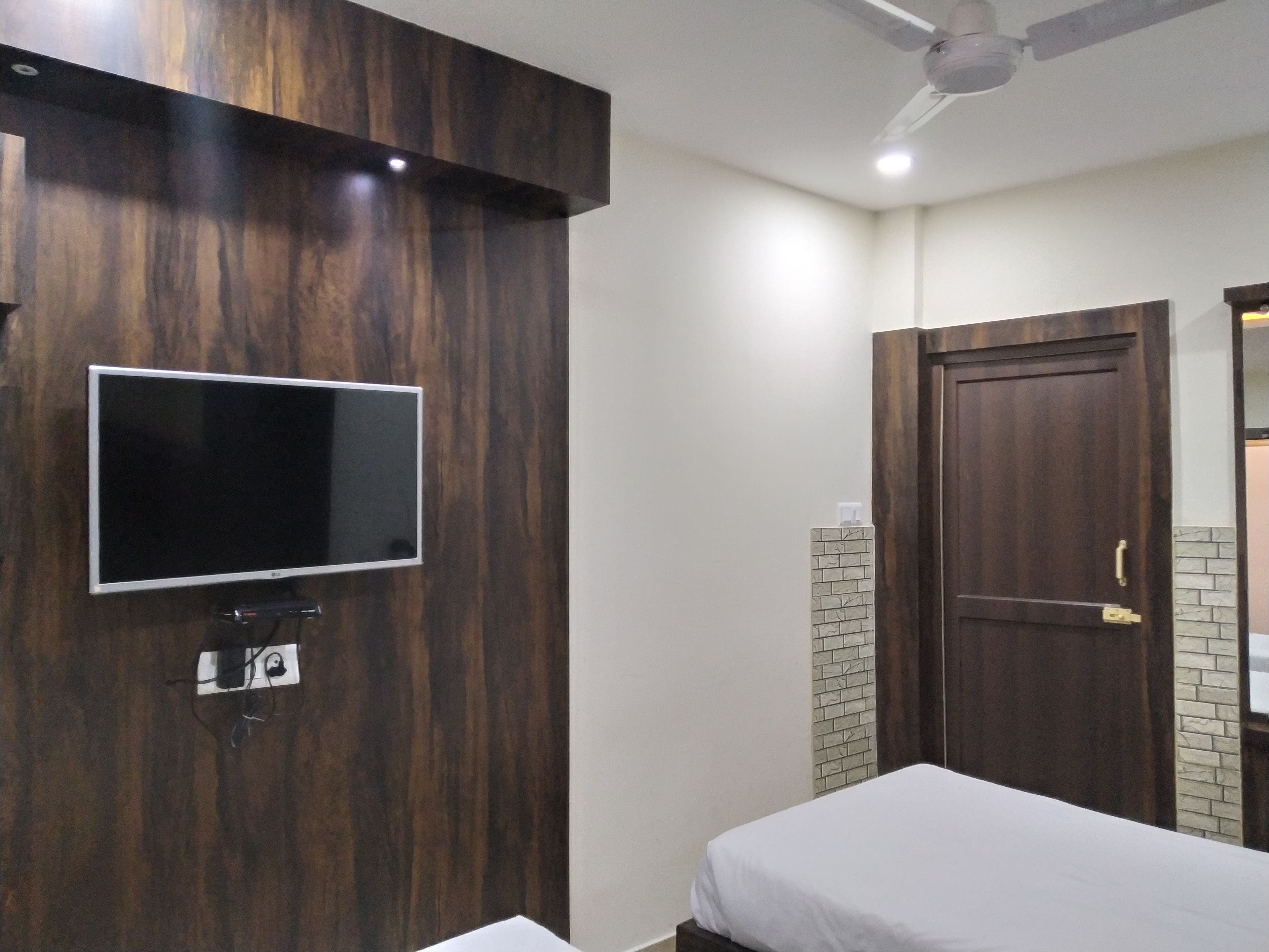 Quarto com televisão fixada numa parede de madeira, cama com roupa de cama branca, porta de madeira e ventoinha no teto.