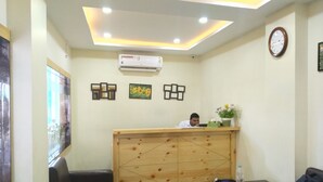 A reception area with a wooden reception desk, a clock on the wall, and a framed picture of sunflowers. - Hotel Bikram (Tezpur)