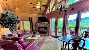 L’intérieur chaleureux d’un chalet avec une cheminée, un plancher en bois, un ventilateur de plafond et de grandes fenêtres offrant une vue panoramique.