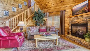 L’intérieur chaleureux d’un chalet avec un escalier en bois, une cheminée, un canapé en cuir et un téléviseur fixé au mur.