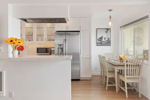 A modern kitchen with white cabinetry, a stainless steel refrigerator, and a dining area with a wooden table and chairs. - Beachfront 2BR Condo in Sunny Carpinteria. Walk Everywhere! (Carpinteria)