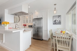 A modern kitchen with white cabinetry, a stainless steel refrigerator, and a dining area with a wooden table and chairs. - Beachfront 2BR Condo in Sunny Carpinteria. Walk Everywhere! (Carpinteria)