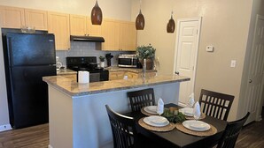 Une cuisine dotée d’un îlot central, de meubles en bois, d’un réfrigérateur noir, d’une cuisinière et d’un four à micro-ondes. On y trouve une table à manger avec des chaises, et une porte blanche en arrière-plan.