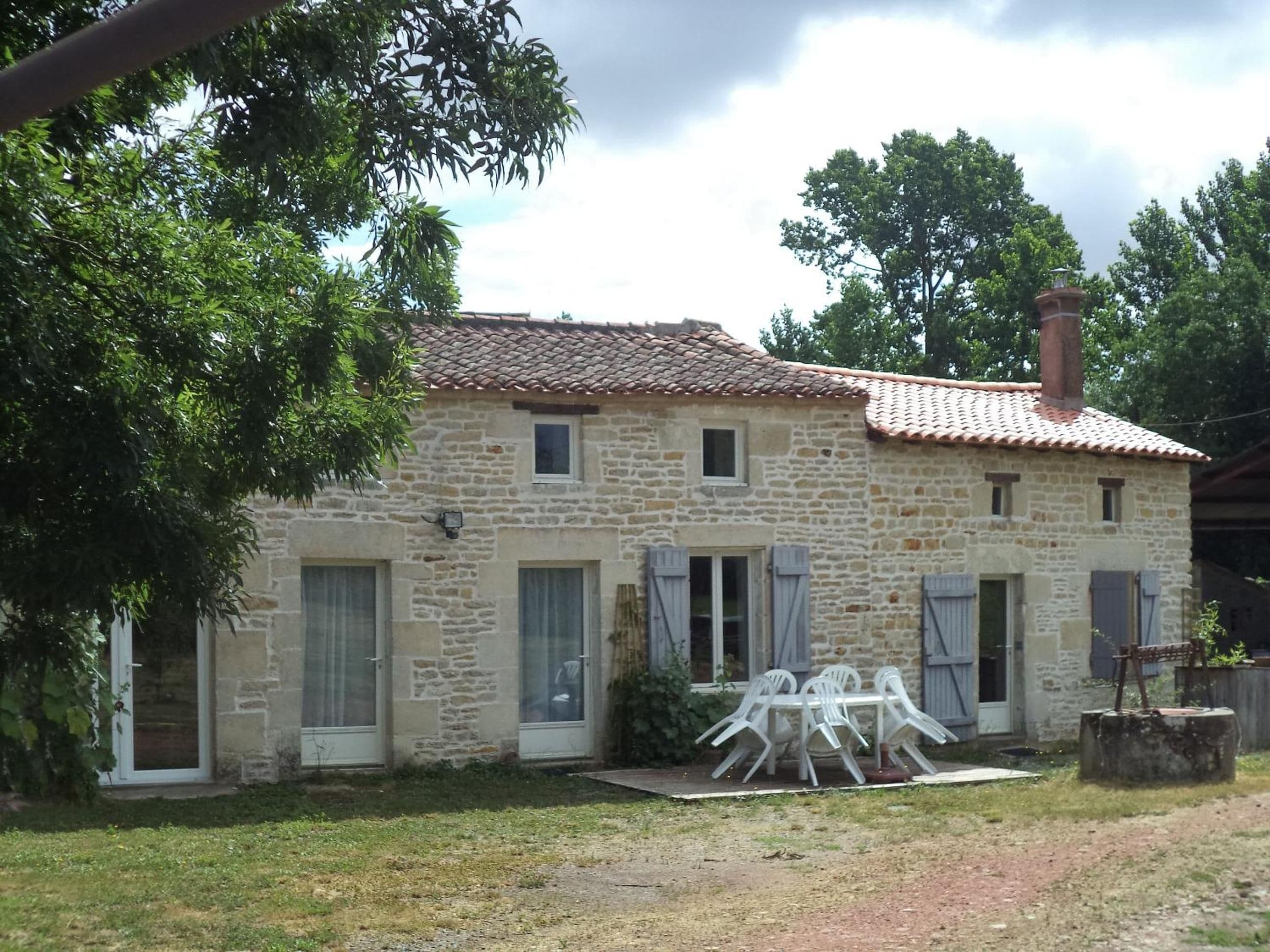 A stone house with a tiled roof, a small porch with plastic chairs, and a chimney.
