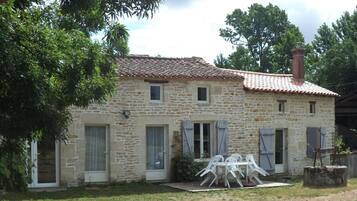 A stone house with a tiled roof, a small porch with plastic chairs, and a chimney.