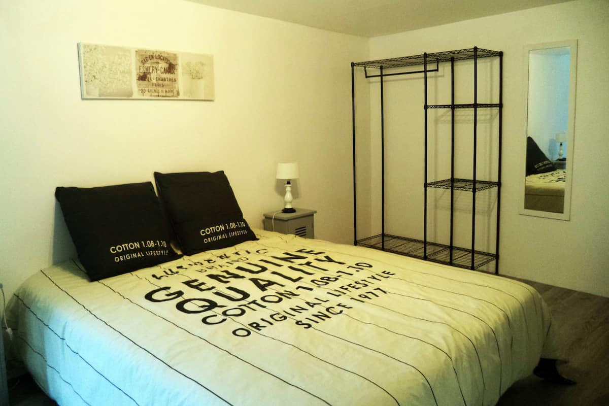 Un dormitorio con cama, dos almohadas, una mesita de noche, una lámpara, un estante metálico negro y un cuadro en la pared.