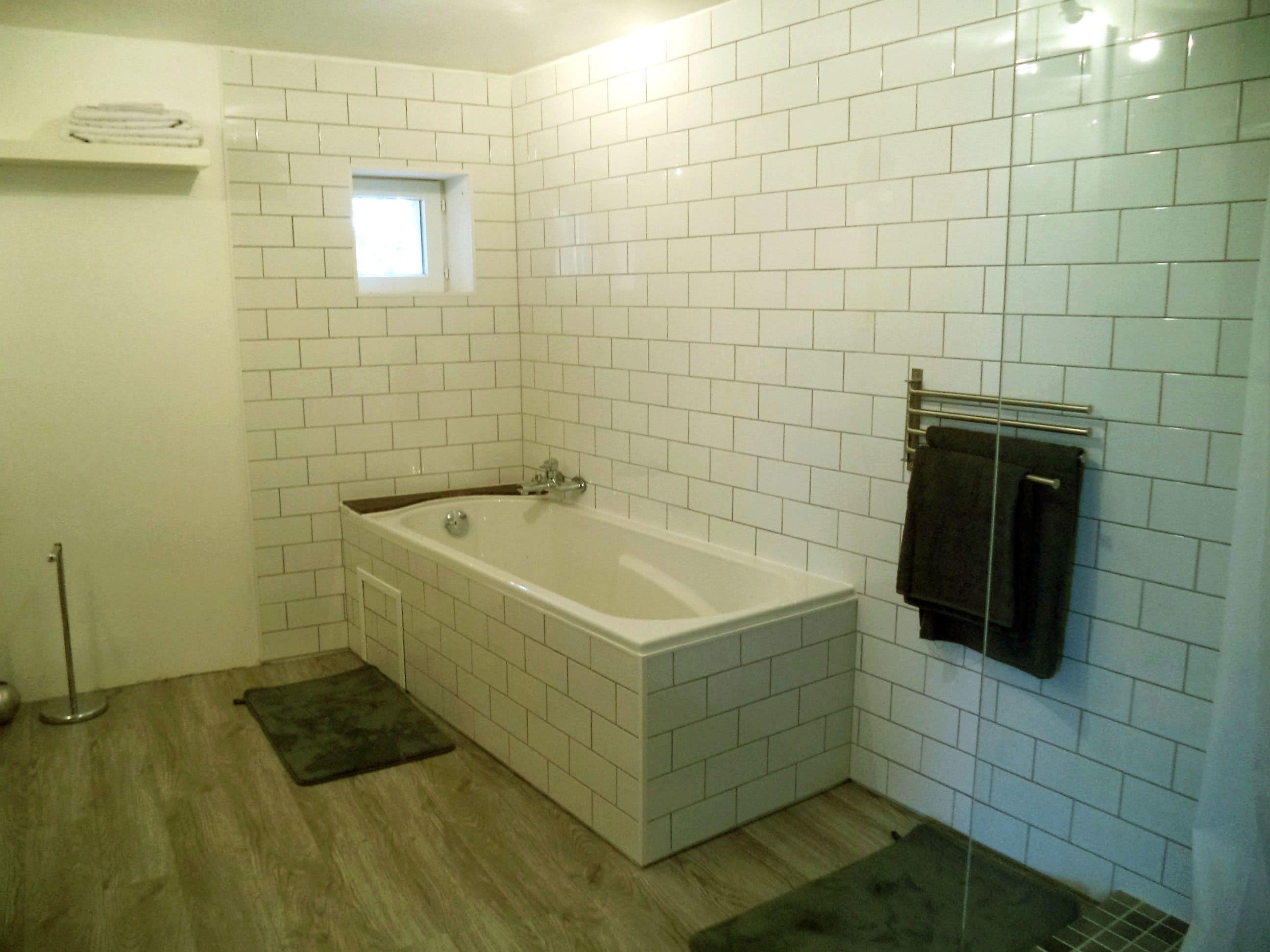 Un baño con bañera, paredes cubiertas de azulejos blancos, una ventana pequeña y un perchero.
