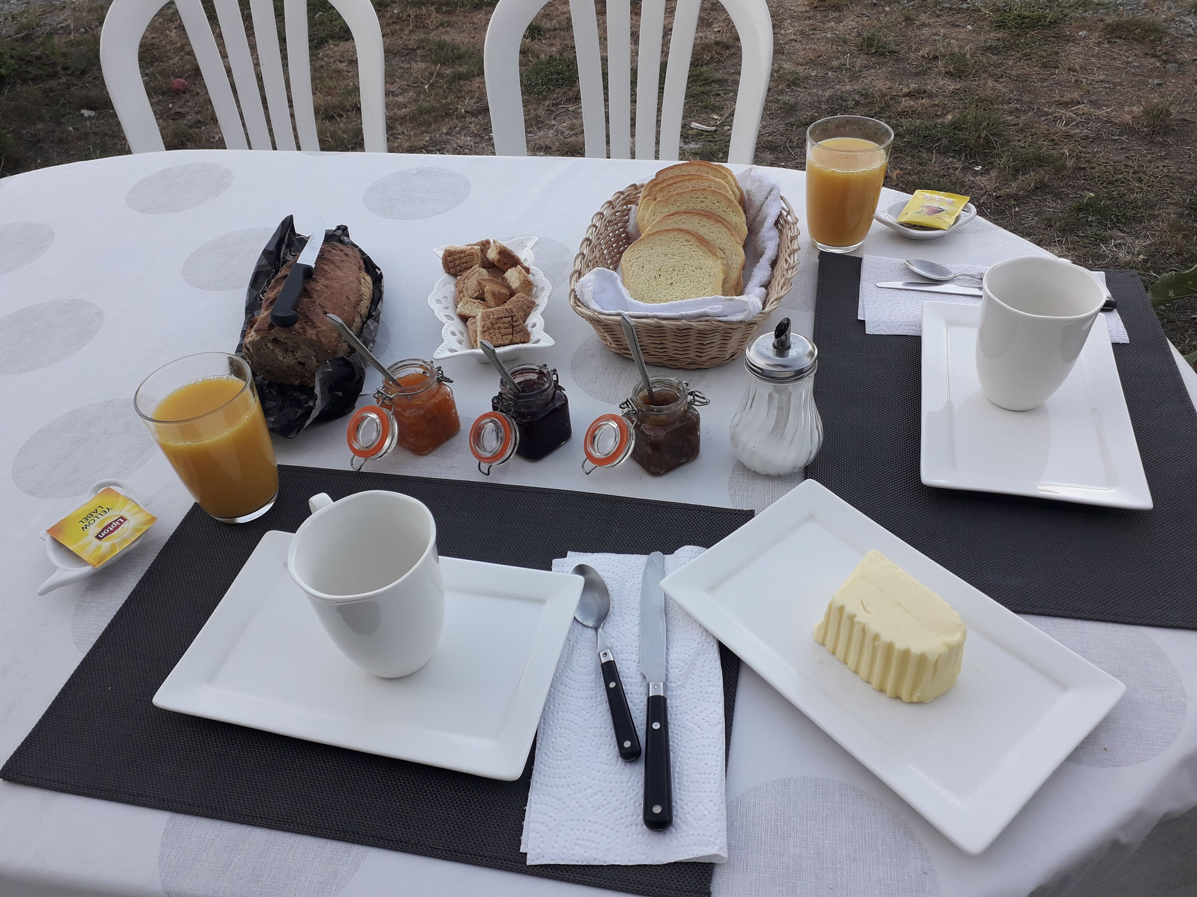 Una mesa de desayuno con pan, mantequilla, mermeladas y un vaso de jugo de naranja.