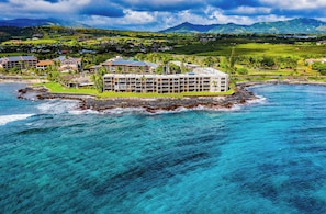 A coastal hotel with multiple buildings, a rocky shoreline, and a view of the ocean and mountains in the background. - End Unit Oceanfront Getaway at Kuhio Shores 401 (Po'ipu)
