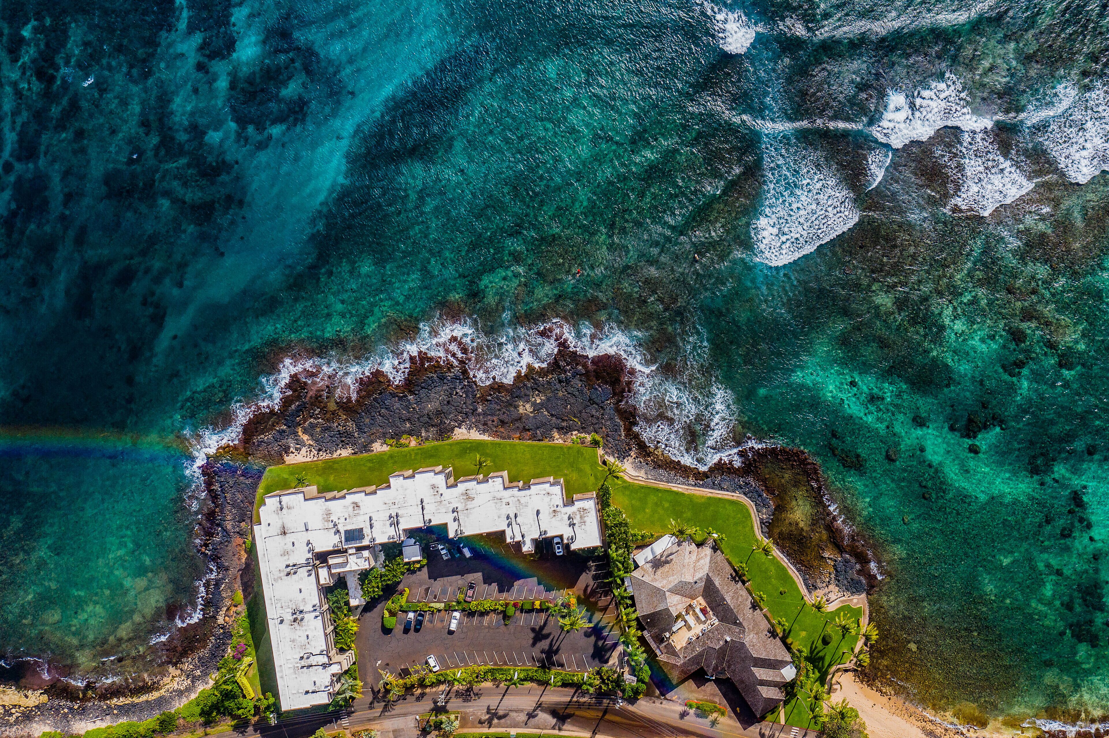 Vista aérea de un hotel frente al mar con estacionamiento, piscina y acceso directo a la playa.