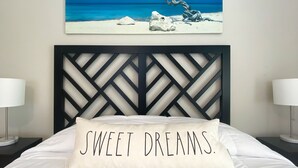 Een slaapkamer met een bed, een kussen met de tekst 'SWEET DREAMS.', een nachtkastje, een lamp en een schilderij van een strand.