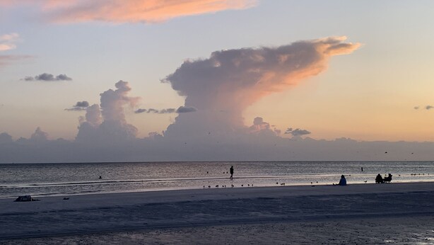 Ein Strand mit einer großen Wolkenformation, ruhiger See und Menschen, die sitzen und spazieren gehen.