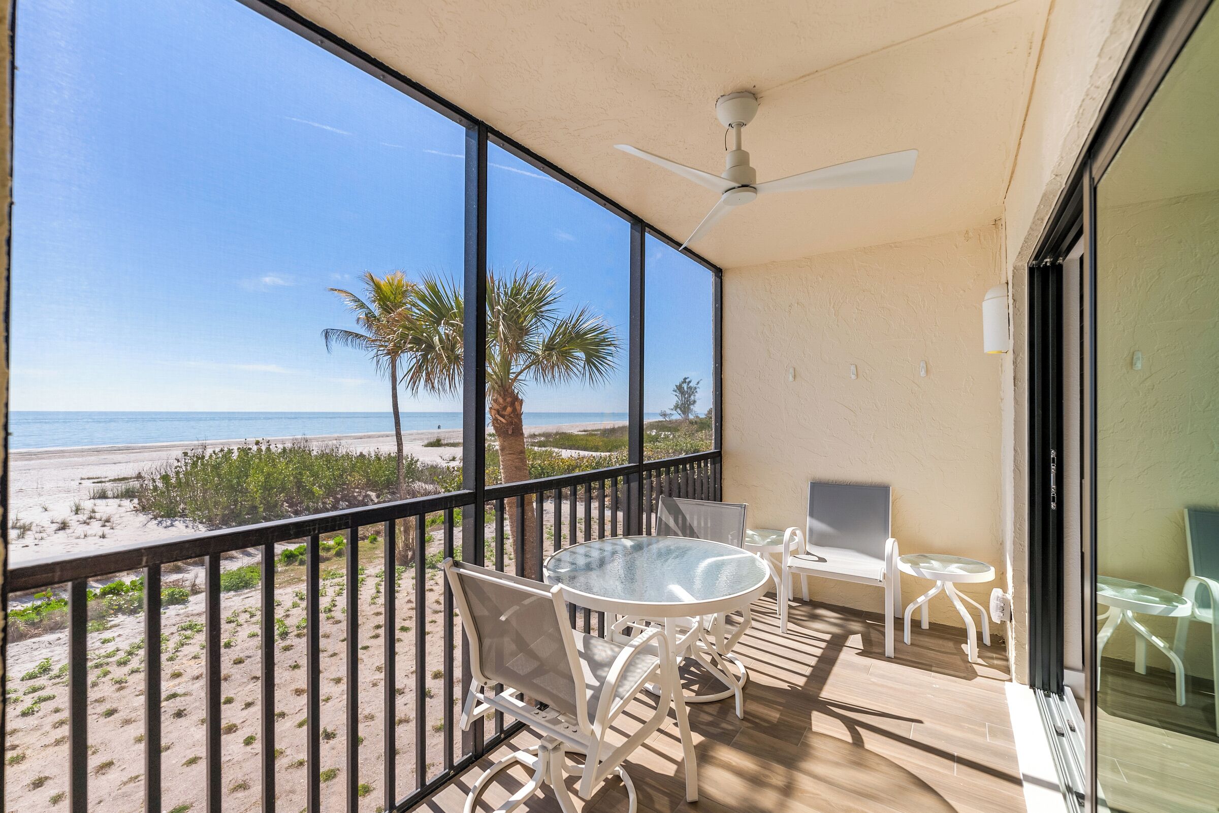 A balcony with a glass table and chairs, a ceiling fan, and a view of the beach and palm trees.