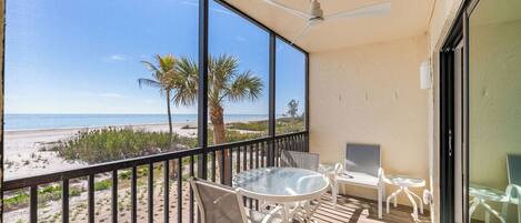 A balcony with a glass table and chairs, a ceiling fan, and a view of the beach and palm trees.